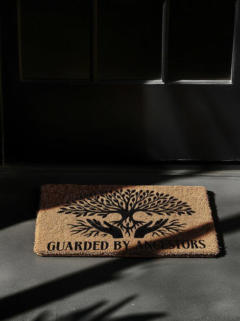 18x24 inch rectangular coir doormat made from natural coconut husk fibers, featuring a bold black print of a stylized tree with branching leaves and intertwined roots forming the shape of protective hands. Below the tree design, the phrase "GUARDED BY ANCESTORS" is printed in large, capitalized serif lettering. The mat has a textured, bristled surface ideal for scraping dirt and moisture from shoes, and a durable construction suited for everyday entryway use. Designed for indoor or outdoor placement, this d